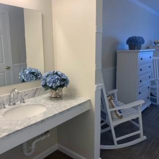 A cozy bathroom with a sink and vanity, complemented by a mirror and decorative flowers; a rocking chair and dresser are visible in the adjoining room.