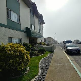 A view of a residential building along a foggy sidewalk with lush greenery and parked vehicles.