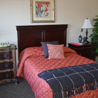 A neatly decorated bedroom with a red quilted bedspread and several pillows, featuring a wooden headboard, artwork on the wall, and a nightstand with a lamp.