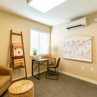 A cozy office space featuring a desk and chair, with natural light from a window, decorated with artwork and a ladder shelf.