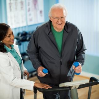 A doctor assists an elderly man during a physical therapy session on a treadmill.