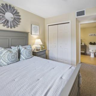 A cozy bedroom featuring neutral tones with a neatly made bed, a nightstand with a lamp, and a closet. The decor includes patterned pillows and a decorative mirror on the wall.