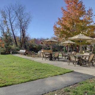 A scenic outdoor patio area with tables, chairs, and umbrellas surrounded by autumn trees and a well-maintained lawn.