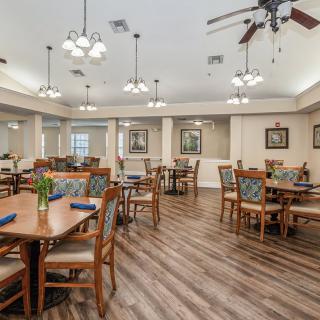 A well-lit dining room with multiple tables and chairs, featuring ceiling fans and chandeliers, and a wood floor.
