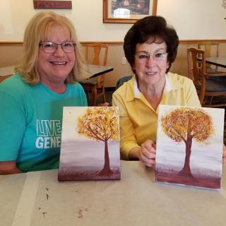 Two women proudly display their autumn tree paintings at an art workshop.