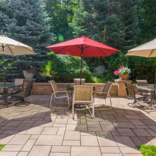 A sunny patio with tables and chairs under colorful umbrellas, surrounded by lush greenery.