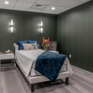 A modern minimalist bedroom with dark green walls, featuring a single bed adorned with blue and white bedding and decorative pillows. A nightstand holds a vase of fresh flowers.