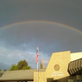 A vibrant rainbow arcs over a building with an American flag flying in front.