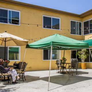 A sunny outdoor patio area with tables, chairs, and canopies in front of a building.