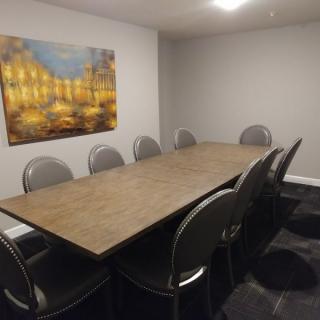 A modern conference room with a large wooden table, surrounded by chairs and featuring a decorative painting on the wall.