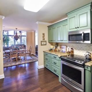 A modern kitchen with green cabinetry opens to an inviting dining area, showcasing hardwood flooring and sleek appliances.