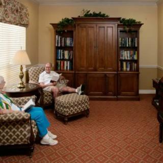 Two elderly individuals sitting in a cozy living room, reading in armchairs beside a large wooden bookshelf.