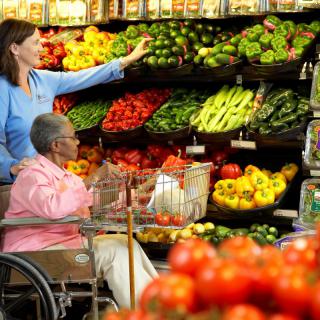 A woman assists an elderly person in a wheelchair while shopping for fresh produce at a grocery store.