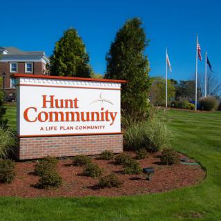 Entrance sign for Hunt Community, a senior living facility, with landscaped surroundings and flags in the background.