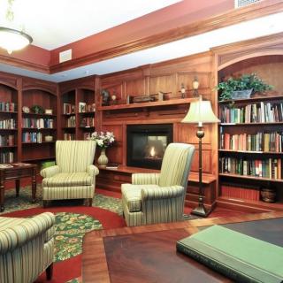 A cozy living room with wood-paneled walls, featuring a fireplace, armchairs, and bookshelves filled with books.