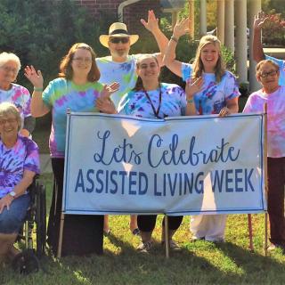 A group of people celebrating Assisted Living Week outdoors with a banner and tie-dye shirts.