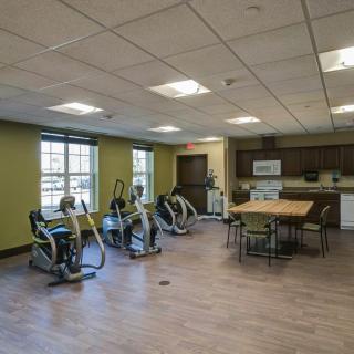 A spacious exercise room features stationary bikes, a kitchen area, and a table with chairs on a wooden floor.