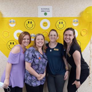 Four women smiling in front of a yellow smiley face backdrop.