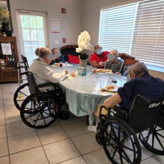 A group of elderly individuals in wheelchairs gather around a dining table in a care home.