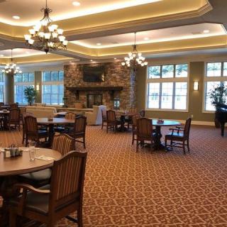 A spacious dining room with elegant decor, featuring multiple tables and chairs, ambient lighting from chandeliers, a cozy fireplace, and large windows allowing natural light.