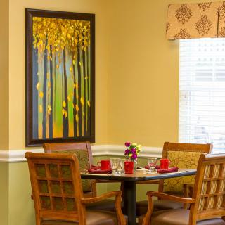 A cozy dining area with a round table, decorated with red cups and small flowers, surrounded by wooden chairs. A vibrant painting of trees adorns the wall.