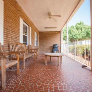 A cozy porch with wooden benches and a table, set against a brick wall and overlooking a fenced garden.