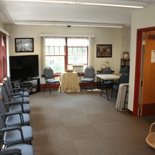 A spacious waiting room with chairs lined up against the walls, a television set, and large windows allowing ample natural light.