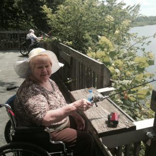 An elderly woman in a wheelchair enjoys a peaceful moment on a wooden deck by the lake, surrounded by lush greenery.