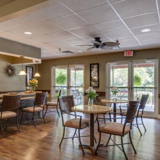 A cozy cafe dining area with tables and chairs, illuminated by natural light through large windows.