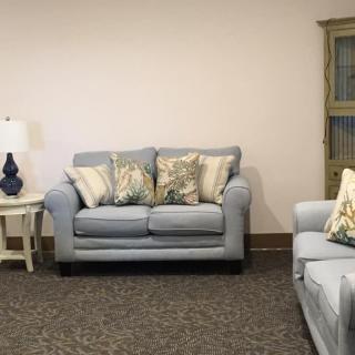 A cozy living room setup featuring a light gray sofa with patterned cushions, a side table with a lamp, and a cabinet in the background.