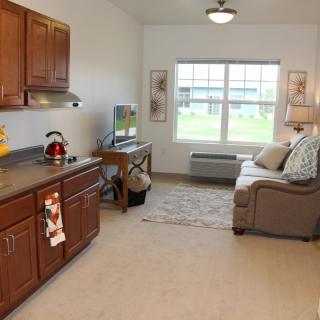 A cozy apartment interior featuring a small kitchen area and a living space with a sofa, TV, and window.