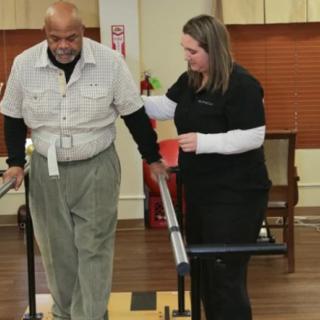 An elderly man receives assistance from a healthcare professional during a physical therapy session using walking aids.