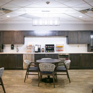 A modern office break room with a central table, chairs, and kitchen amenities.