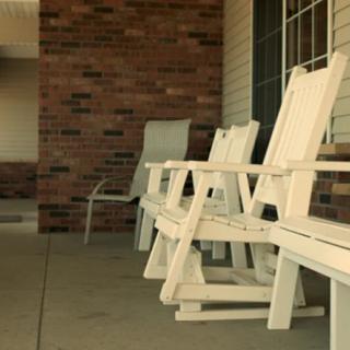 A row of white rocking chairs sits on a covered porch with a brick wall backdrop.