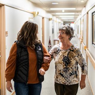 A caregiver assists an elderly woman as they walk together down a hallway.