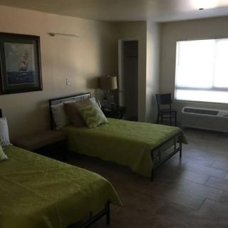 A bedroom with two beds featuring green bedding, a nightstand with a lamp, and a window letting in natural light.