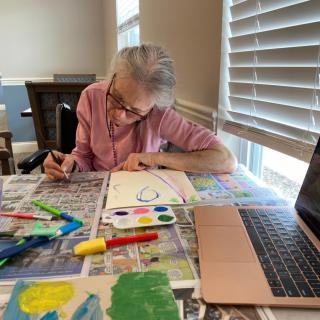 An older adult engaged in painting at a table, surrounded by art supplies and using a laptop for reference.