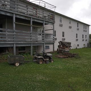 An outdoor view showcasing a multi-unit building with a porch, a quad bike, and stacked firewood on the lawn.