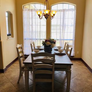 A cozy dining room with a wooden table set for four, illuminated by a chandelier and natural light from large windows.