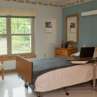 A minimalist room with a hospital bed, natural light streaming in through a large window, and simple furnishings.