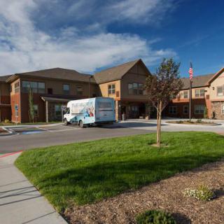 A moving truck is parked in front of a large residential building surrounded by a landscaped lawn on a partly cloudy day.