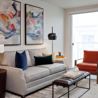 A modern living room with a grey sofa, vibrant artwork, and a cozy orange chair.
