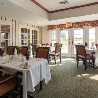 A well-lit dining area with neatly arranged tables and chairs, featuring tablecloths and natural light streaming through the windows.