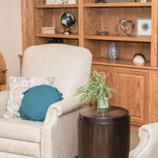 A cozy living room corner features a comfortable armchair with a blue pillow, a small round side table with a plant, and a wooden bookshelf filled with decorative items.