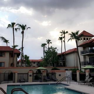 A scenic view of a resort featuring a swimming pool, surrounded by buildings and palm trees under a cloudy sky.