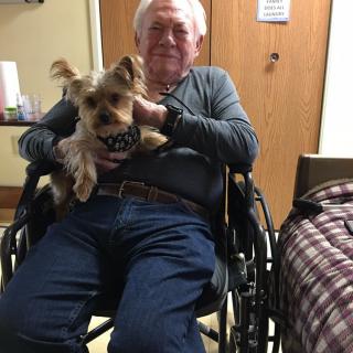 An elderly man sitting in a wheelchair holding a small dog indoors.