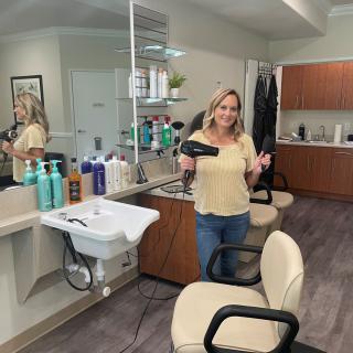 A woman stands in a hair salon holding a blow dryer, surrounded by hair care products.