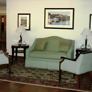 A cozy living room with elegant furniture, including a sofa, armchairs, and coffee tables adorned with lamps. Artwork hangs on the wall above a patterned rug.