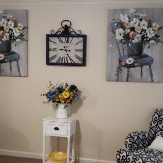A cozy interior scene featuring floral wall art, a decorative clock, and a vase of flowers on a white side table.