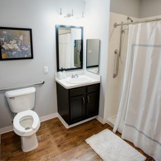 A modern bathroom with a toilet, sink, mirror, and shower curtain on wooden flooring.